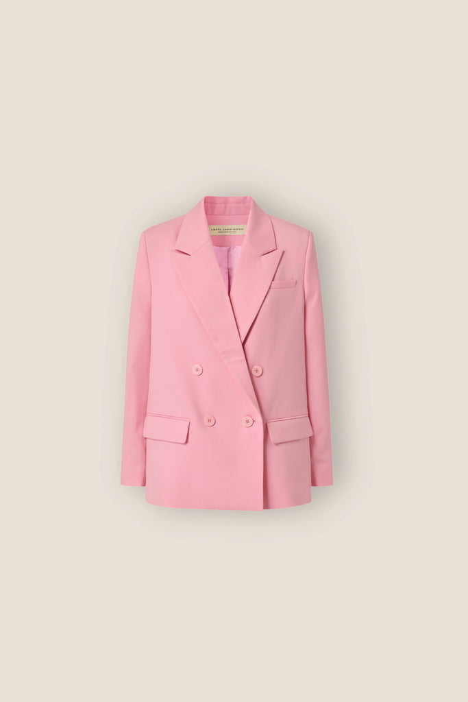 The ALVA Blazer, a candy pink double-breasted piece with notched lapels and a slightly oversized fit, is displayed against a plain beige background. Embracing sustainable fashion, it features two side flap pockets and a chest welt pocket.