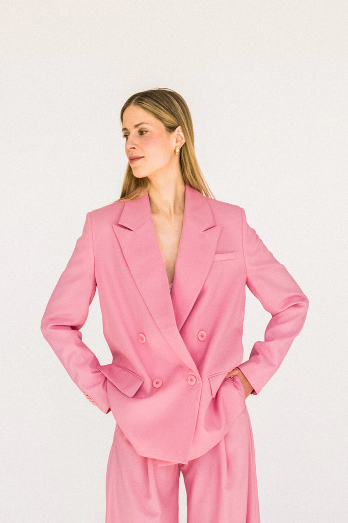 A person with long blond hair stands against a plain white background, wearing the ALVA Blazer in Candy Pink and matching pants. They look to the side with hands on hips, exuding confidence in their sustainable suit.