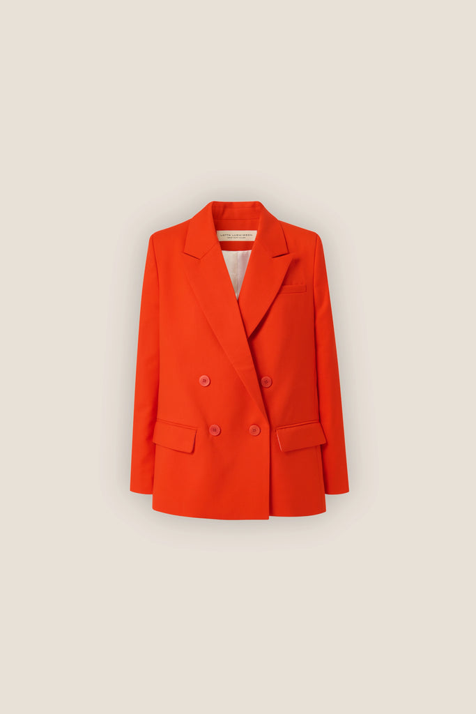 The ALVA Blazer, a tangerine red double-breasted piece with notched lapels and a slightly oversized fit, is displayed against a plain beige background. Embracing sustainable fashion, it features two side flap pockets and a chest welt pocket