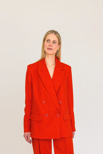 A person with long blond hair stands against a plain white background, wearing the ALVA Blazer in Tangerine Red and matching pants. They look to the side with hands on hips, exuding confidence in their sustainable suit.