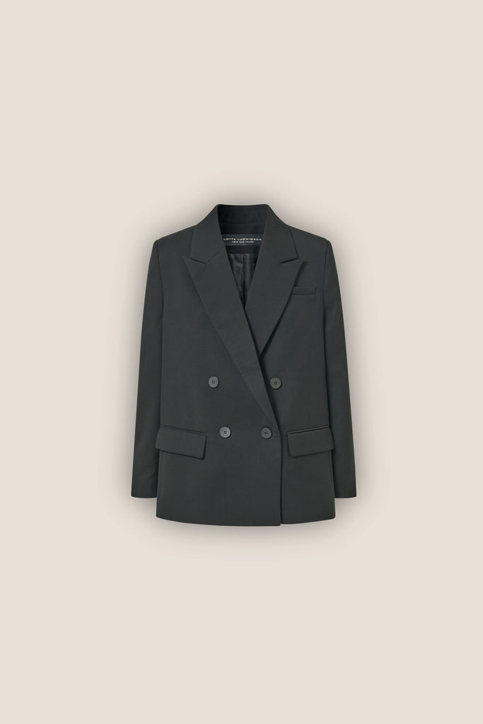 The ALVA Blazer, a tuxedo black double-breasted sustainable piece with notched lapels and a slightly oversized fit, is displayed against a plain beige background.