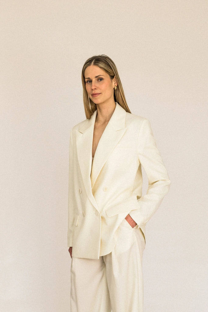 A person with long blond hair stands against a plain white background, wearing the ALVA Blazer in Vanilla Creme and matching pants.